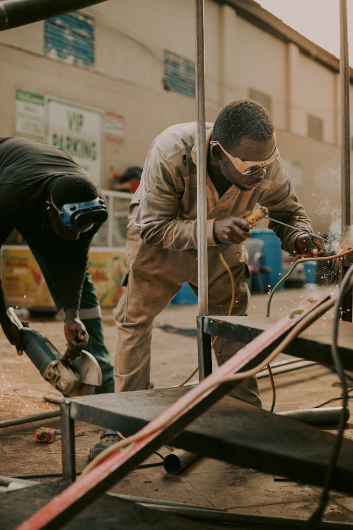 Two male workers wearing safety gear welding metal outdoors, showcasing teamwork and industrial labor.