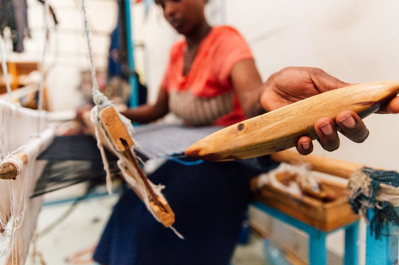 Side view of crop unrecognizable African American craftswoman in casual wear holding shuttle with weft thread while working on wooden weaving loom machine in studio